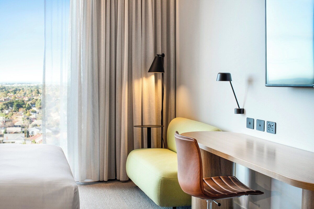A hotel room with a bed, a desk, a chair, a lamp, and a view of a cityscape.