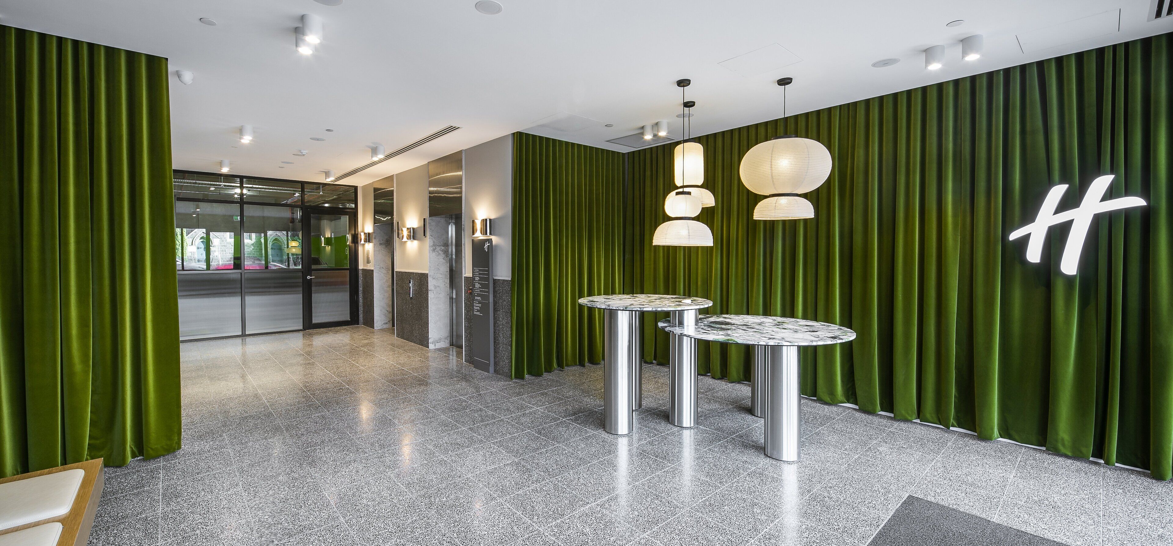 A modern hotel lobby with green curtains, marble-topped tables, and a prominent 'H' logo on the wall.