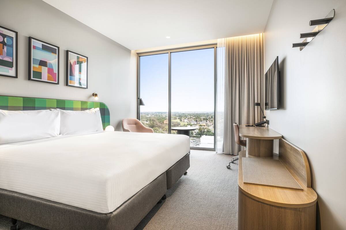 Una habitación de hotel moderna con una cama grande, un escritorio, una silla, un televisor y vistas a la ciudad.