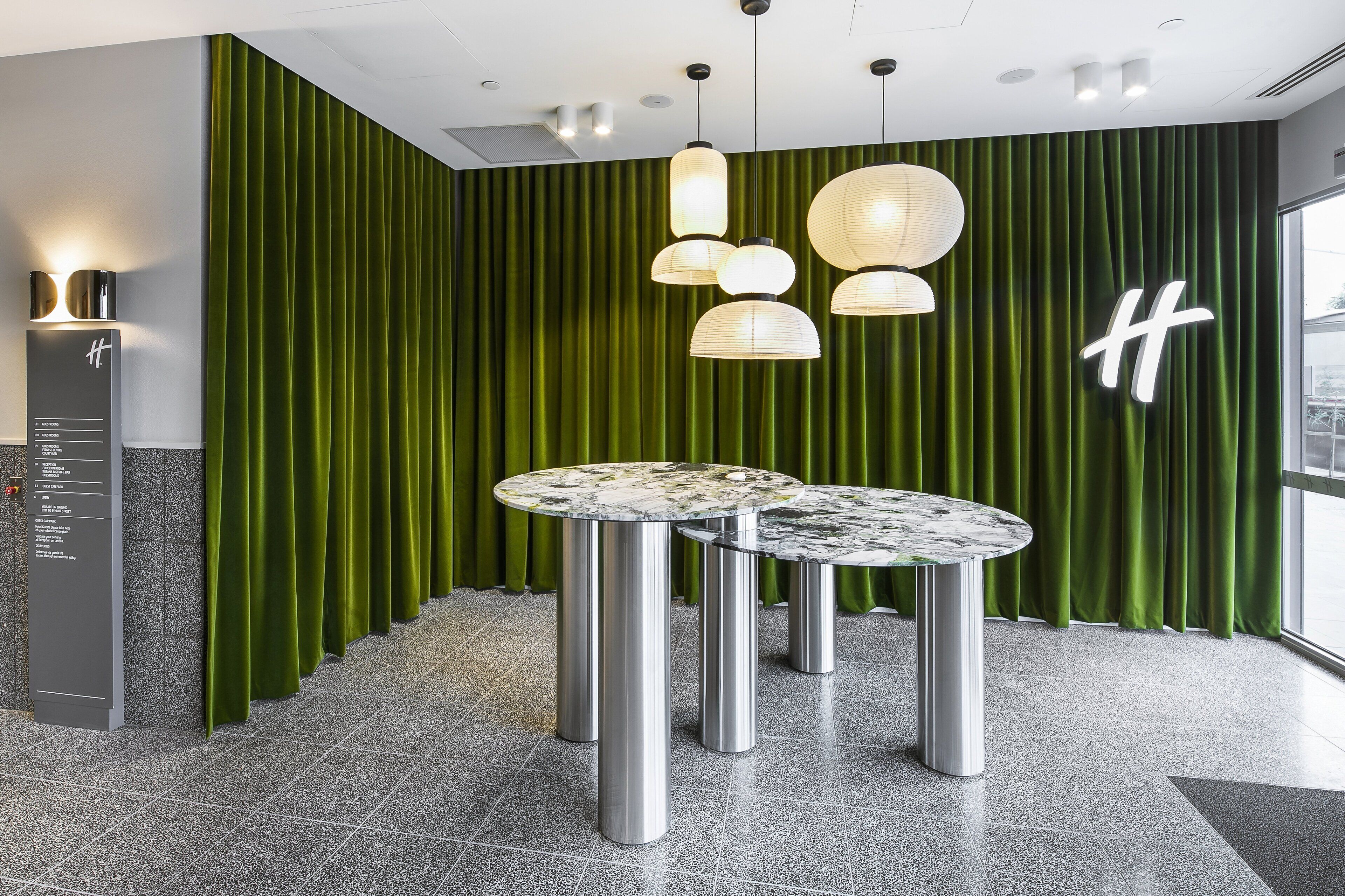 A modern lobby with green curtains, a marble table, and a 'H' sign on the wall.