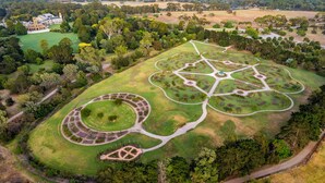Vista aérea de uma grande propriedade com um jardim circular central, um edifício semelhante a um castelo e áreas ajardinadas ao redor.