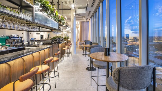 Uma área de bar moderna com banquetas altas, um balcão com copos e taças, e vista para a cidade através de grandes janelas.
