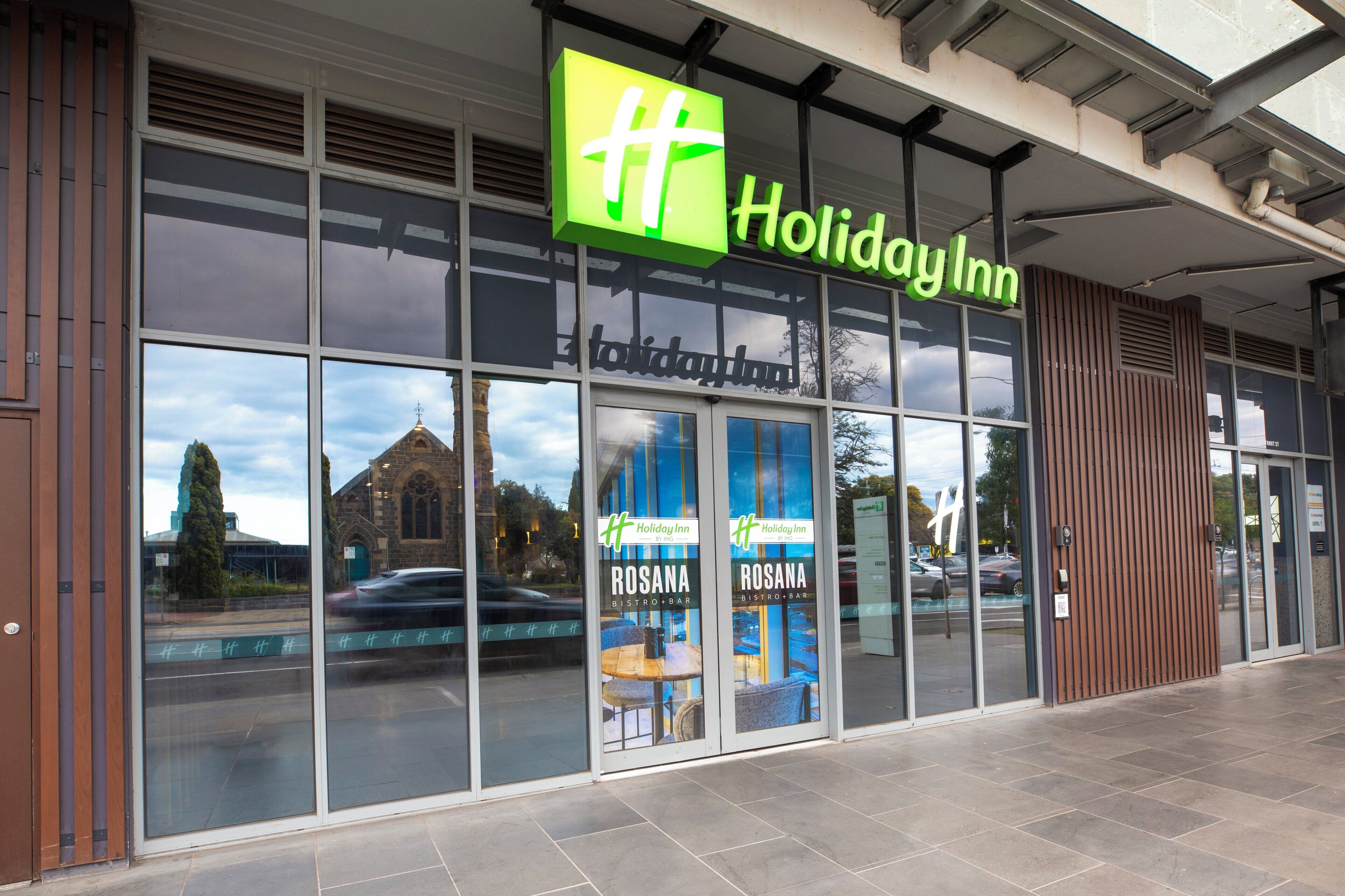 Ein Holiday Inn mit einer großen Glasfassade, einem sichtbaren Außenbereich zum Sitzen und einem Kirchenspiegelbild in den Fenstern.