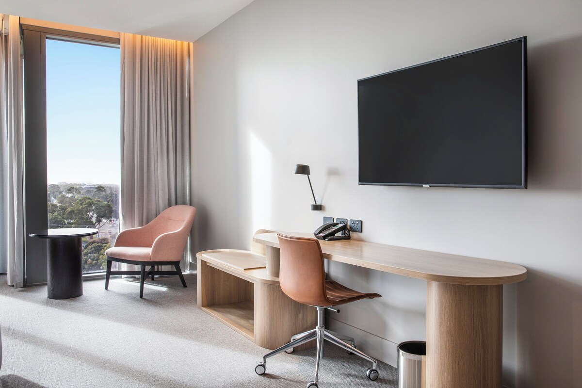 A modern hotel room with a large window, a flat-screen TV mounted on the wall, a desk with two chairs, and a telephone.