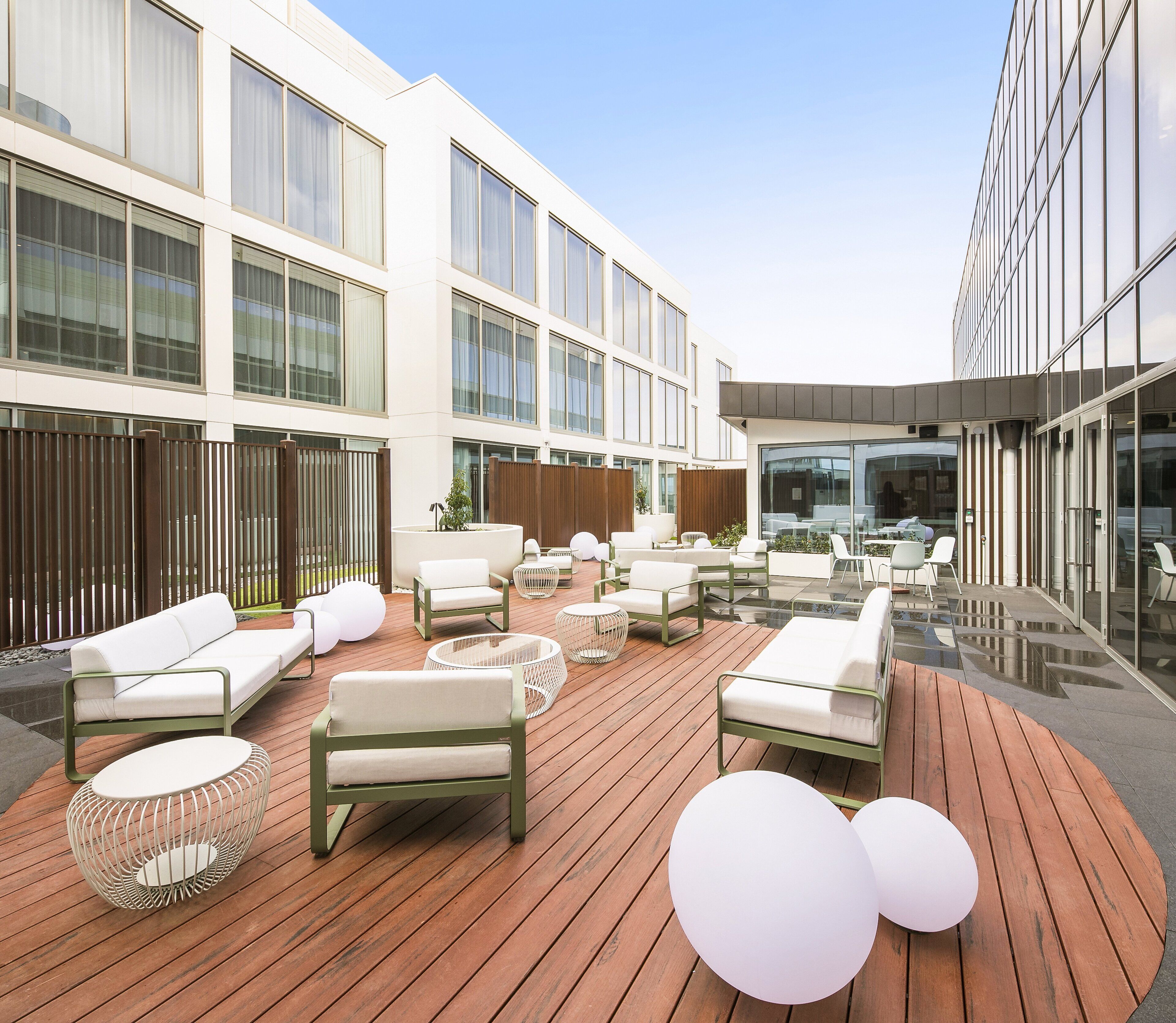 A modern outdoor seating area with sofas and round tables, surrounded by a wooden deck and glass walls of a contemporary building.
