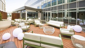 Uma área externa moderna com assentos, incluindo vários sofás e cadeiras brancas, um deck de madeira e grandes janelas refletindo o céu.