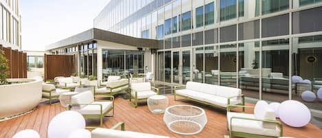 Un área de estar exterior moderna con varios sofás y sillones blancos, una terraza de madera y amplios ventanales que reflejan el cielo.
