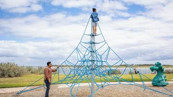 Una persona parada en una escalera de cuerda unida a una estructura de red azul en un entorno exterior, con un cuerpo de agua y una estatua de dragón al fondo.