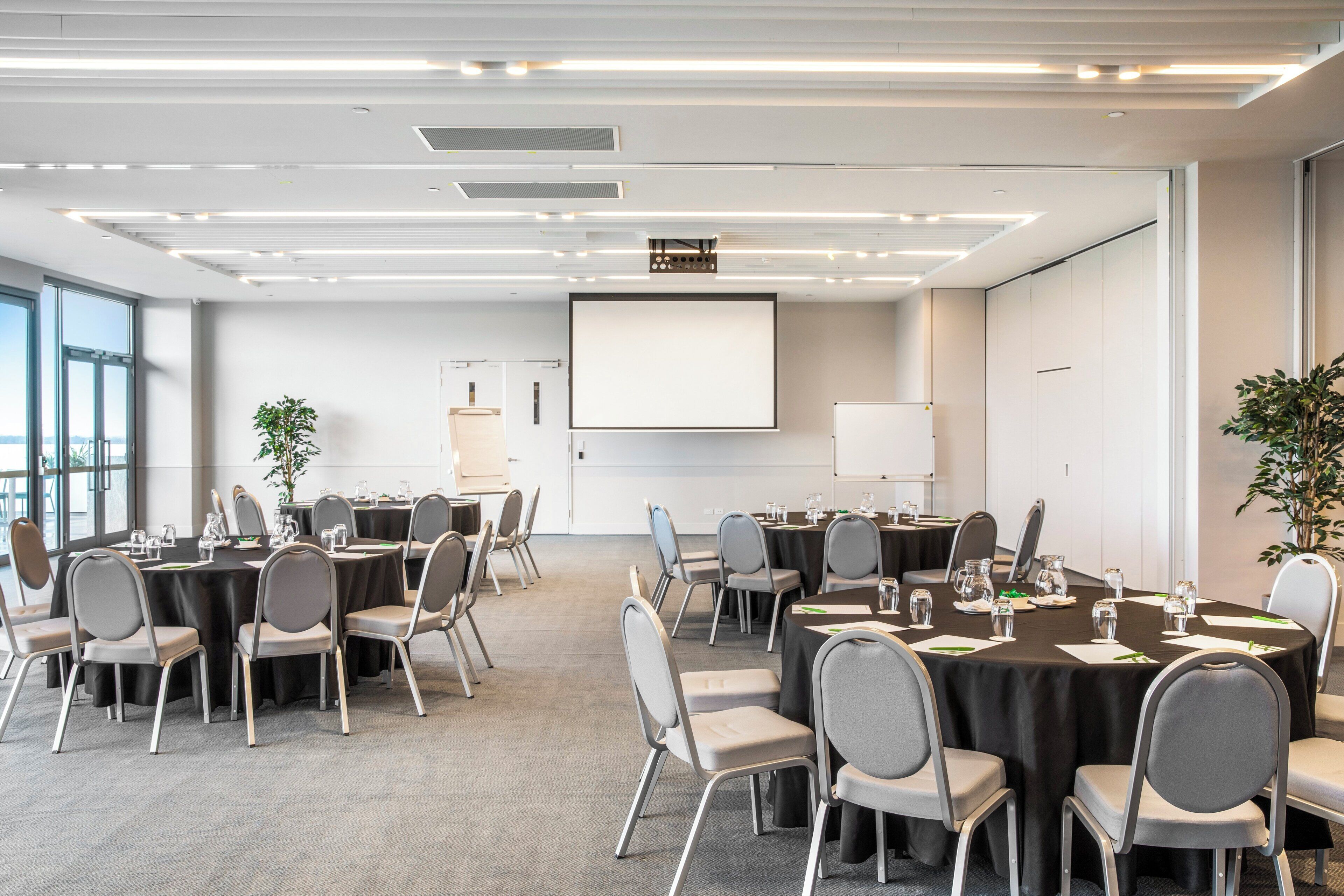 Une salle de conférence avec des tables rondes, des chaises grises et un grand écran.