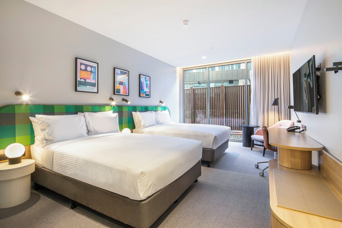 A hotel room with two beds, a desk, a chair, a TV, and artwork on the wall.
