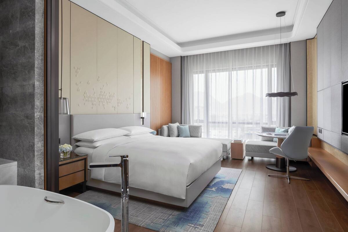 A modern hotel room with a large bed, a sitting area with a sofa and armchair, a flat-screen TV, and a large window with curtains.