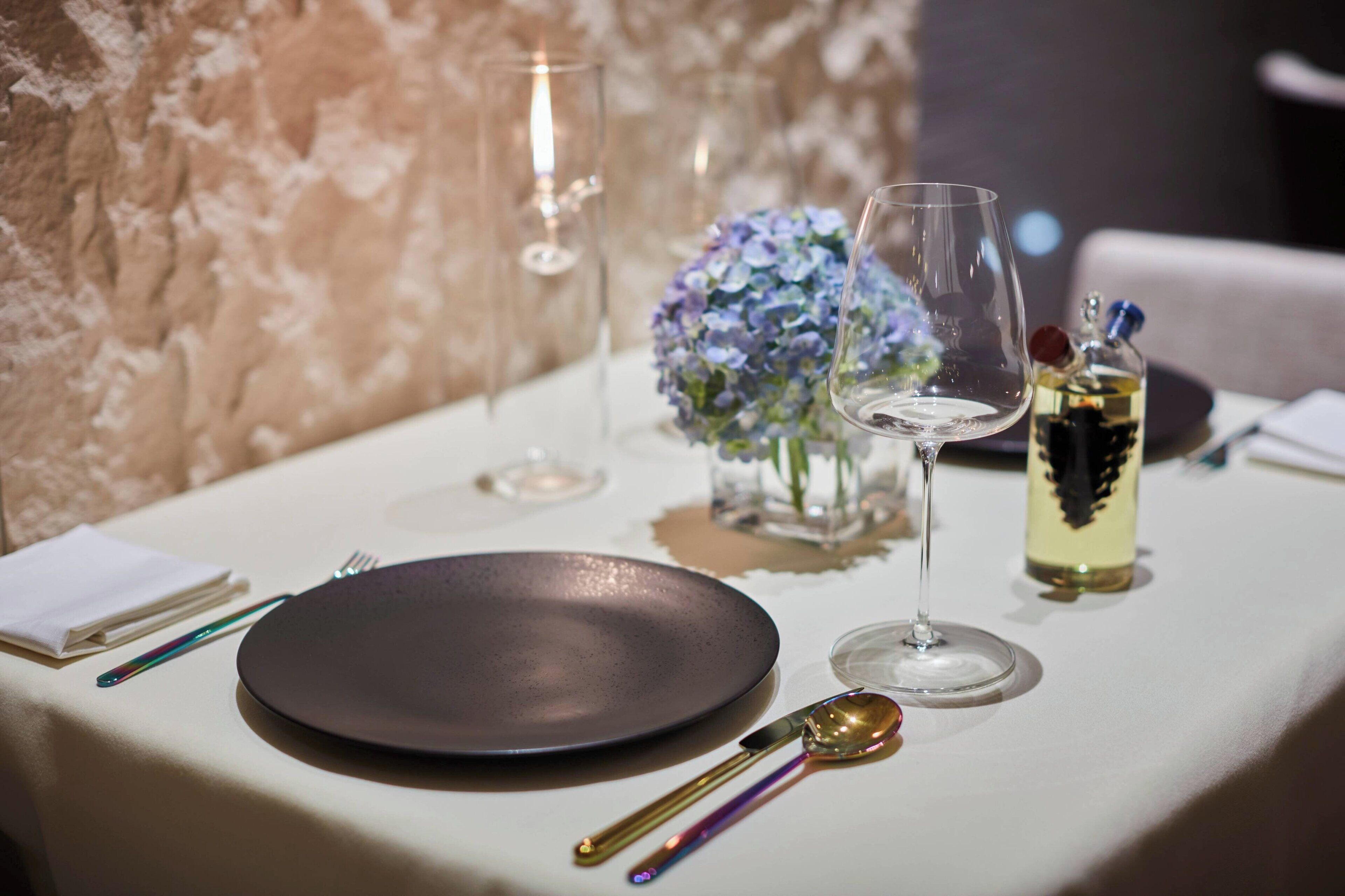 A table set with a dark plate, a spoon, a glass, a bottle of white wine, and a vase with blue flowers.