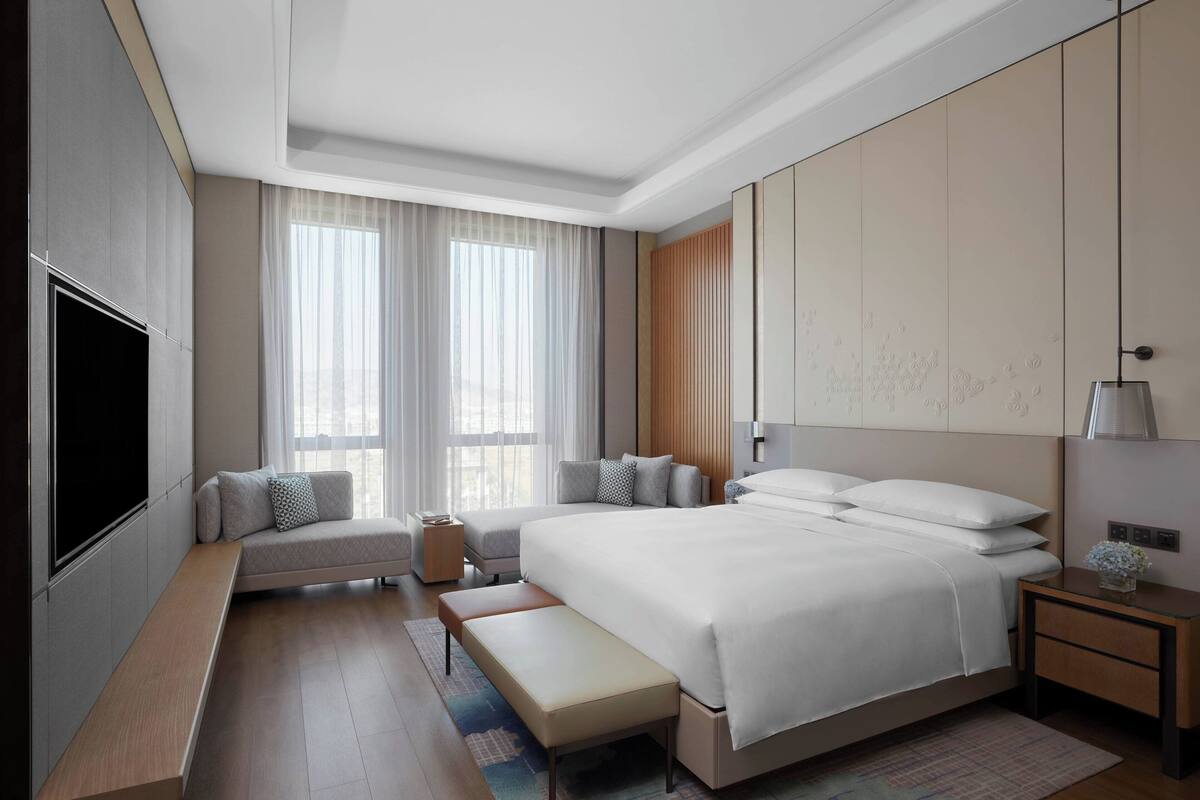 A modern hotel room with a large bed, a bench, a TV, and a bedside table with a lamp.
