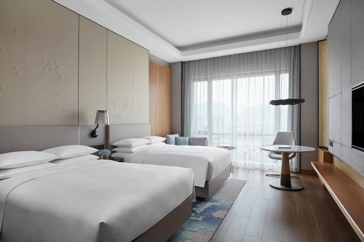 A modern hotel room with two beds, a dining table, a TV, and a large window with curtains.