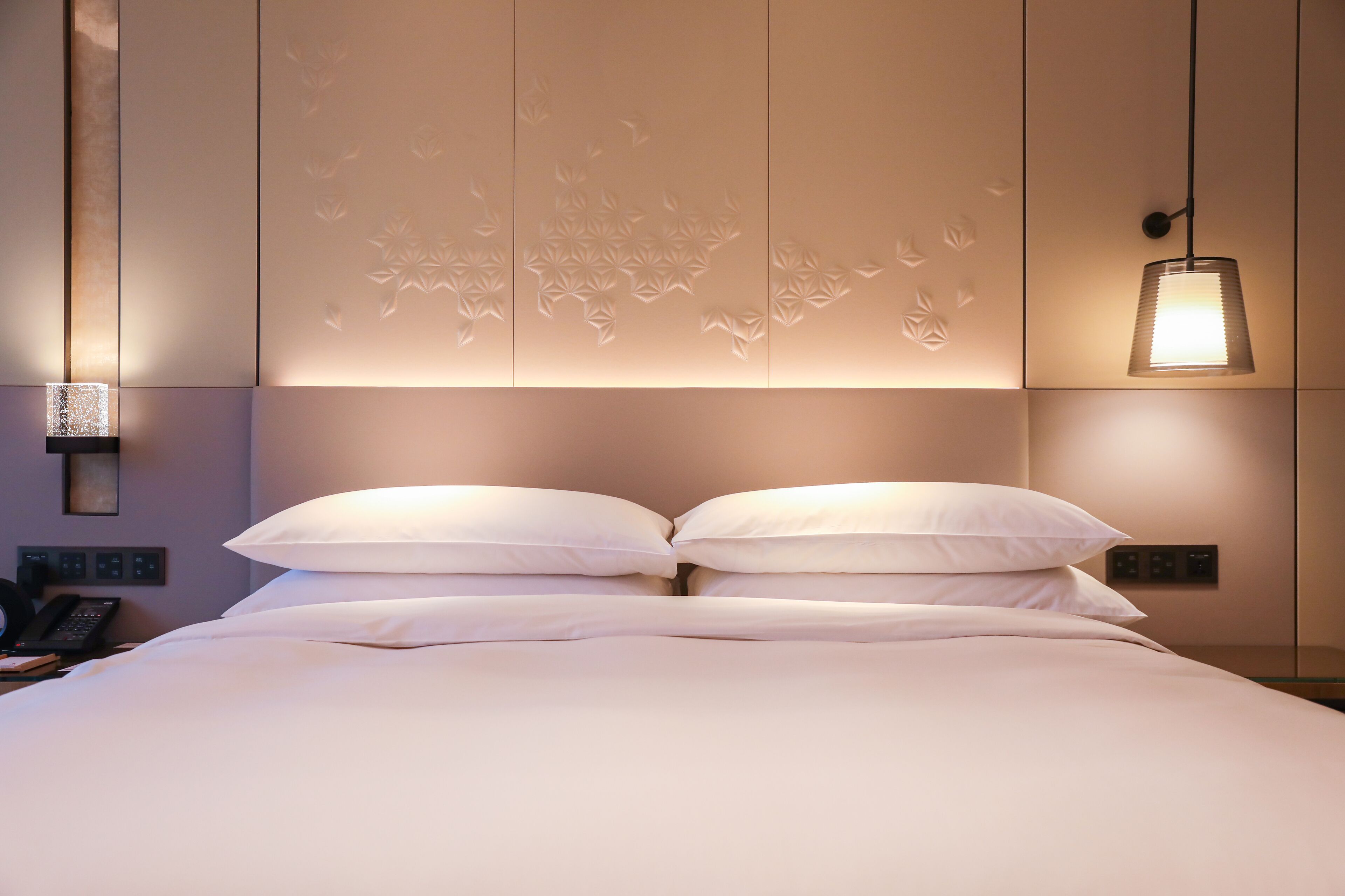 A neatly made bed with white linens and pillows, a wall-mounted phone, and a hanging light fixture.