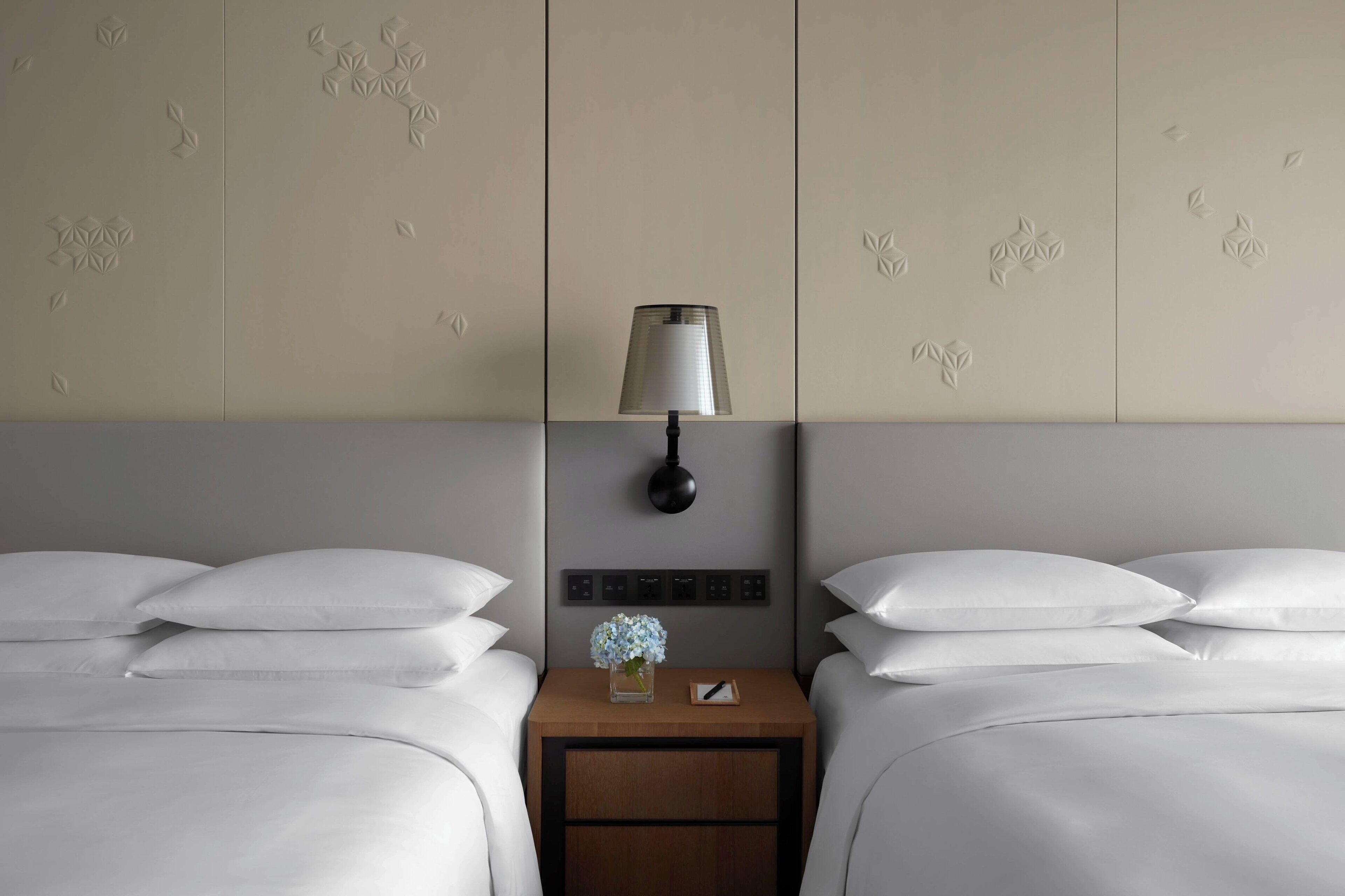 Une chambre d’hôtel avec deux lits, une table de chevet avec une lampe et un panneau de commande fixé au mur.