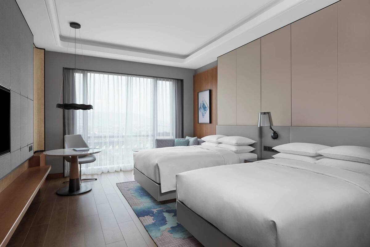A modern hotel room with two beds, a desk, and a large window with sheer curtains.