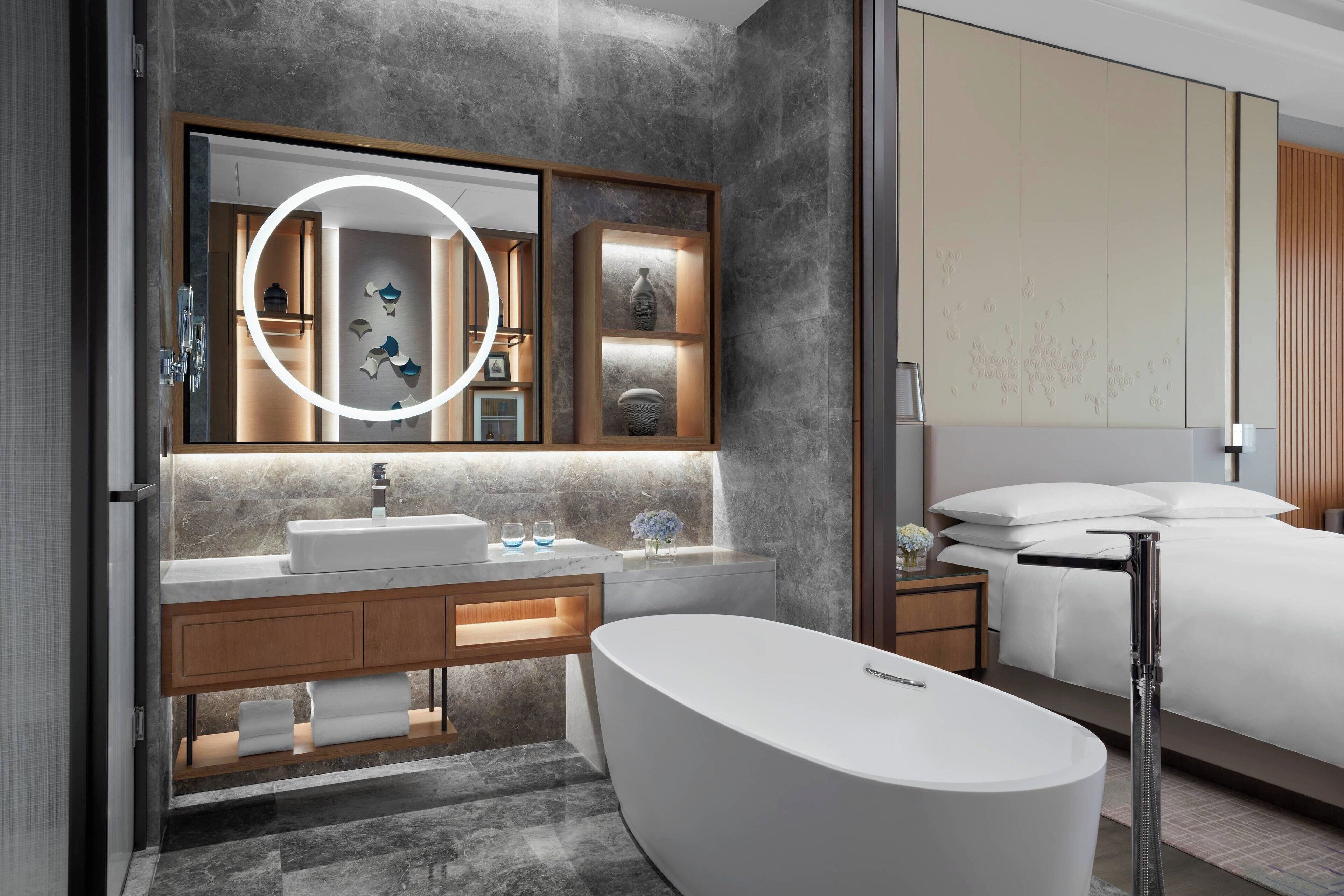 A modern bathroom with a freestanding bathtub, a large mirror with integrated lighting, a vanity with a sink, and a bedside table with a lamp.