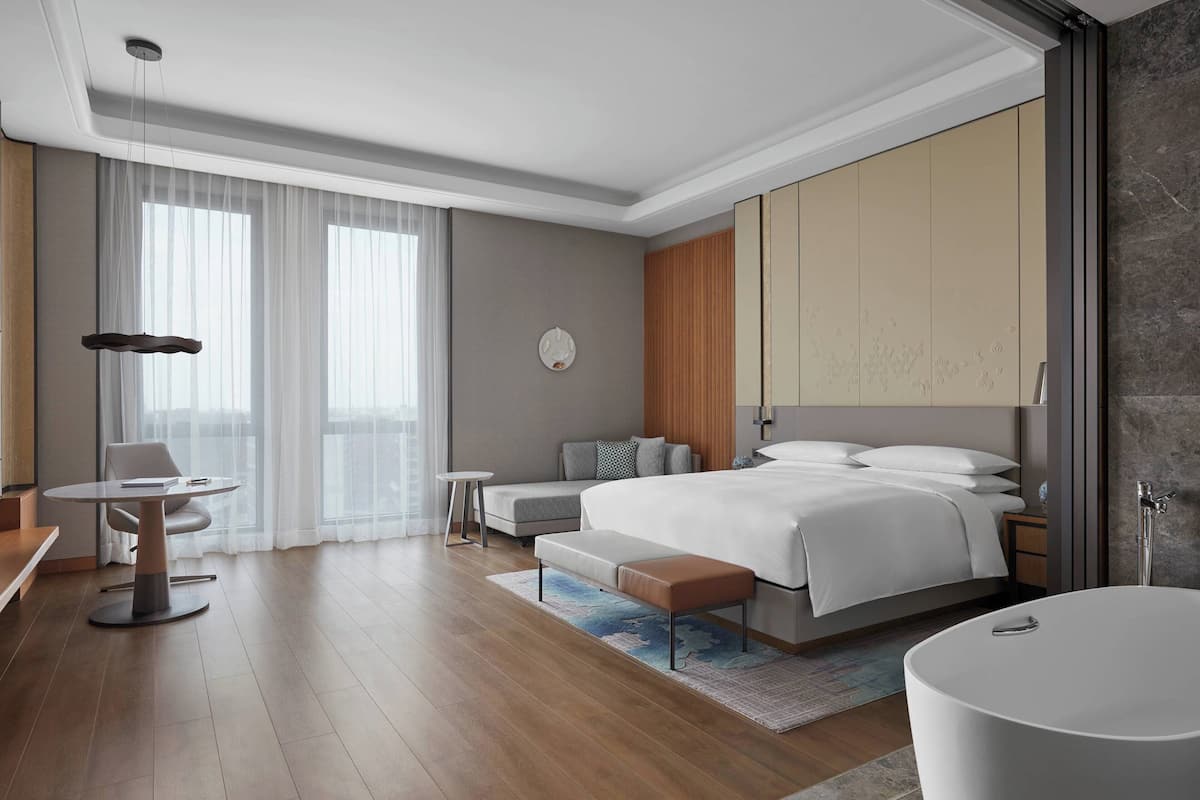 A modern hotel room with a large bed, a sitting area with a table and chairs, a small desk, a sofa, and a bathroom with a bathtub.