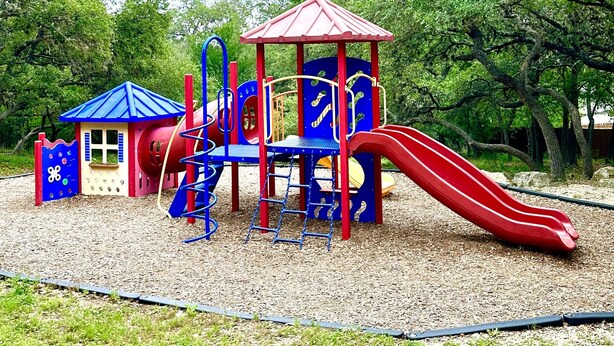 Ein Spielplatz mit einer roten Rutsche, Klettergerüsten und einem kleinen, blau und rot gestrichenen Gebäude.
