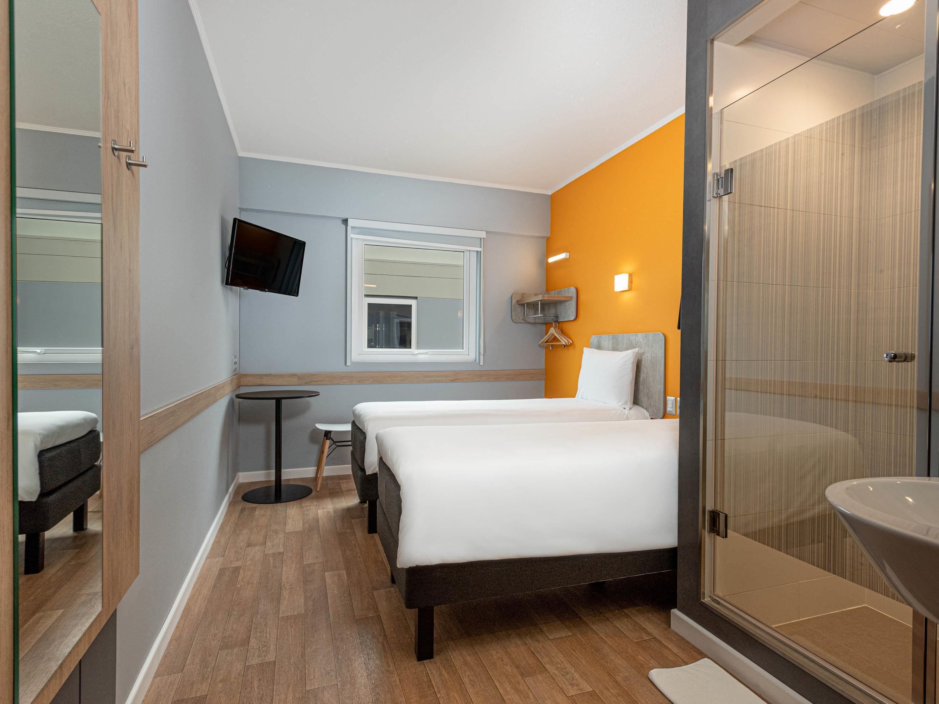 A hotel room with a bed, a small table, a TV mounted on the wall, and a bathroom with a glass shower enclosure.