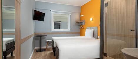 A hotel room with a bed, a small table, a TV mounted on the wall, and a bathroom with a glass shower enclosure.