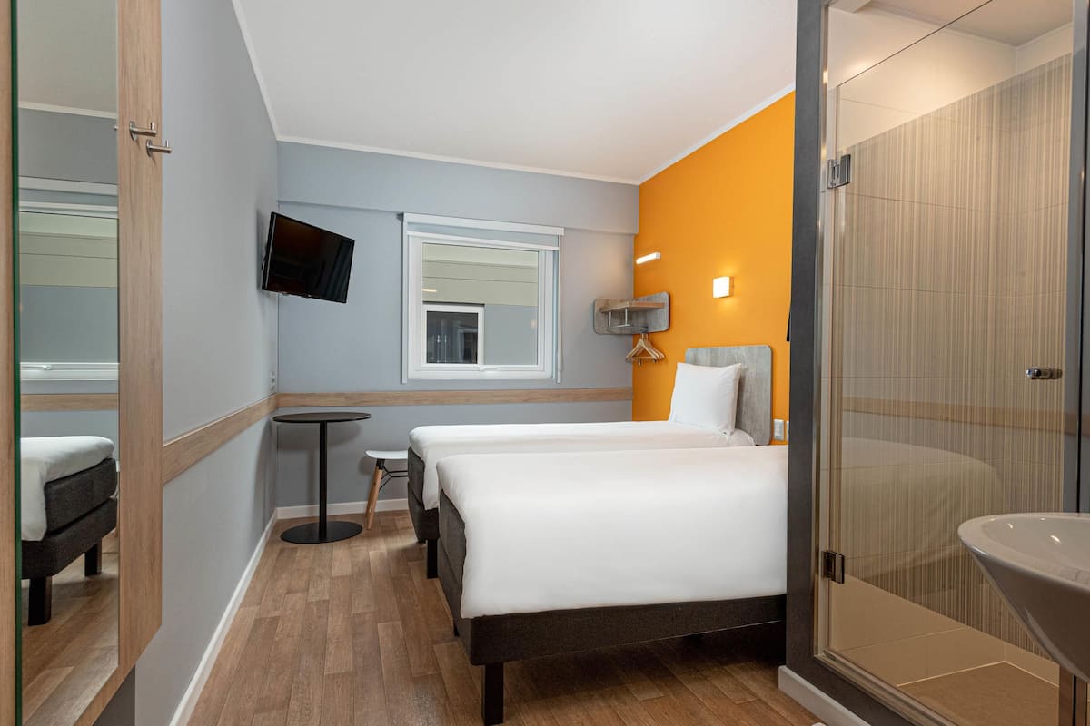 Quarto de hotel com cama, uma mesinha, televisão e banheiro com chuveiro de vidro.