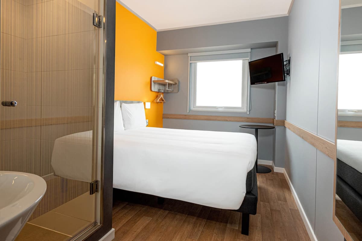 Quarto de hotel com cama, mesa de cabeceira, televisão, janela e chuveiro.