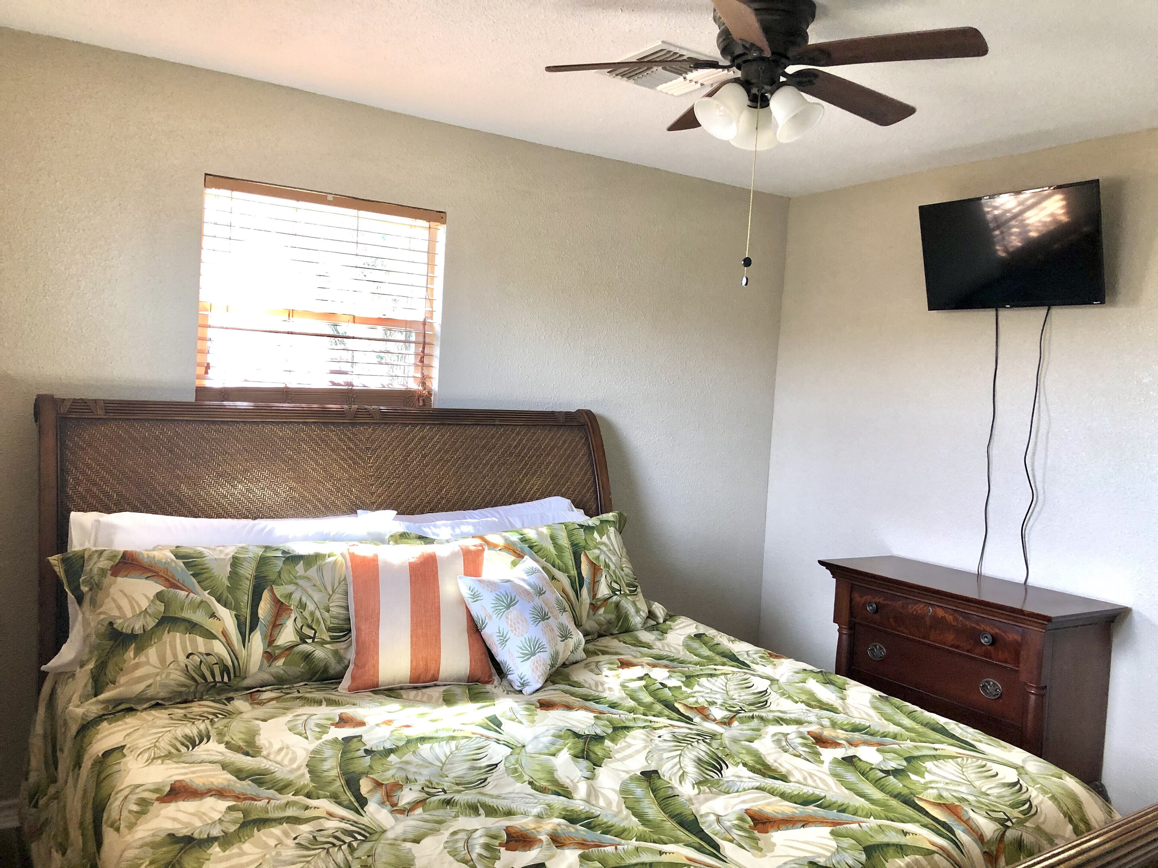A bedroom with a bed, a ceiling fan, a window, and a TV mounted on the wall.
