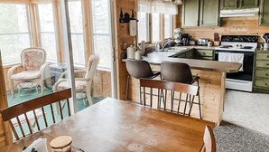 A cabin interior with a wooden dining table, two chairs, a kitchen with a stove and sink, and a window with a view of the outdoors.