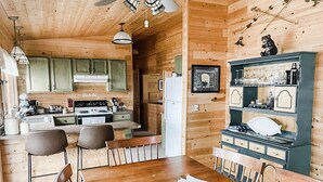 A wooden cabin interior with a dining table, chairs, a kitchen with wooden cabinets, and a shelf with glassware.