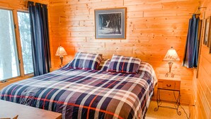 A cozy wooden cabin bedroom with a large bed, two bedside tables with lamps, a framed picture on the wall, and a window with curtains.