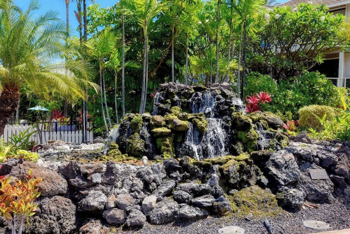 A waterfall feature with surrounding tropical vegetation and a white picket fence.