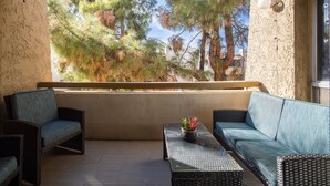 Een balkon met rieten meubels, een glazen tafel en uitzicht op bomen en gebouwen.