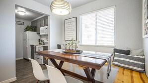 Een moderne keuken met witte kasten, een magnetron en een koelkast. Er staat een eettafel met stoelen en een bank met gestreepte kussens. Een raam met jaloezieën is zichtbaar, en een decoratief bordje hangt aan de muur.