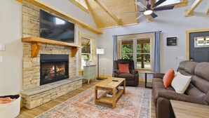 A spacious living room with a stone fireplace, a ceiling fan, and large windows. - New Log Cabin in the Valley, Elk Experience, Year Round Access (Maggie Valley)
