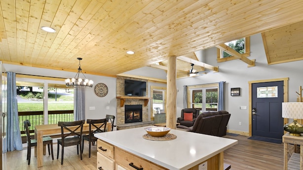A spacious living area with a wooden ceiling, a fireplace, a dining table with chairs, and a large window overlooking a garden. - New Log Cabin in the Valley, Elk Experience, Year Round Access (Maggie Valley)