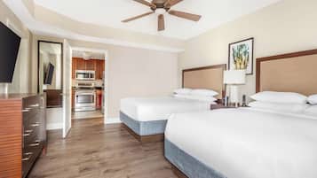 A hotel room with two beds, a ceiling fan, a TV, a wooden dresser, and a kitchenette in the background.