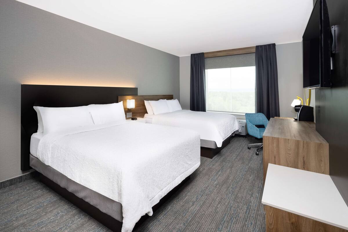 A hotel room with two beds, a desk, a chair, a TV, and a window with curtains.