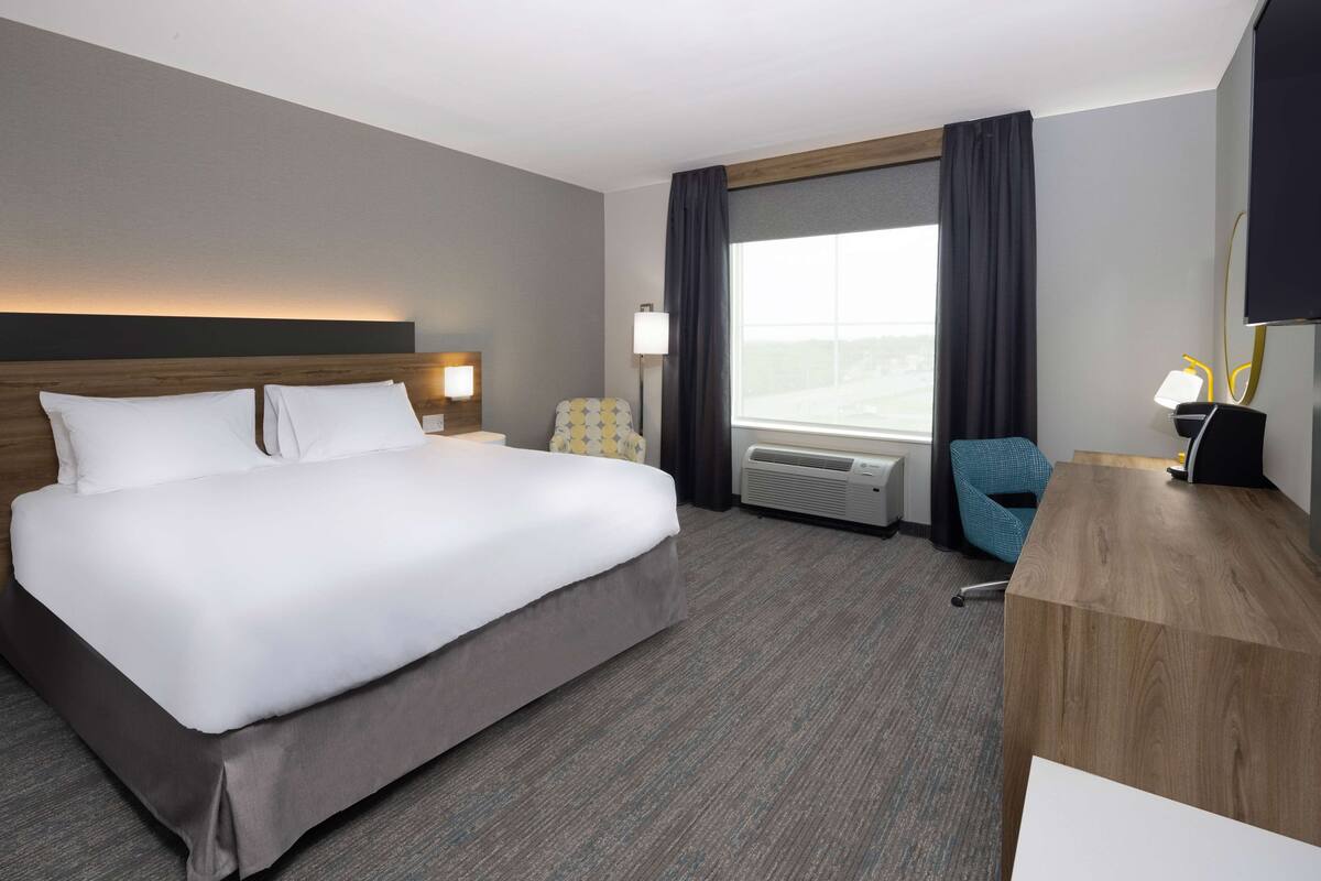 A hotel room with a large bed, a desk, a chair, a desk lamp, a window with curtains, and an air conditioning unit.