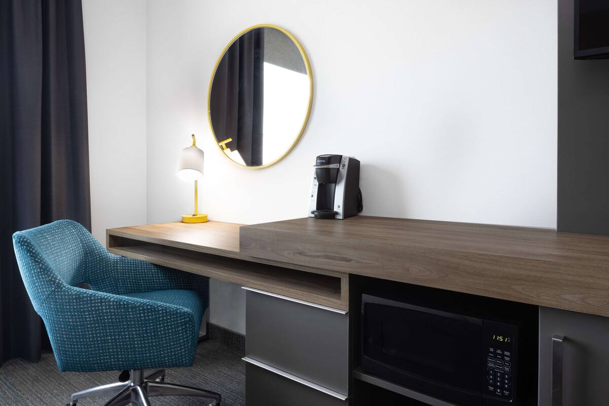 A hotel room with a wooden desk, a blue chair, a round mirror, a lamp, a coffee maker, and a microwave.