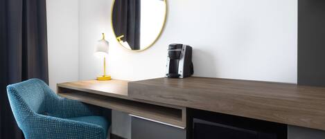A hotel room with a wooden desk, a blue chair, a round mirror, a lamp, a coffee maker, and a microwave.