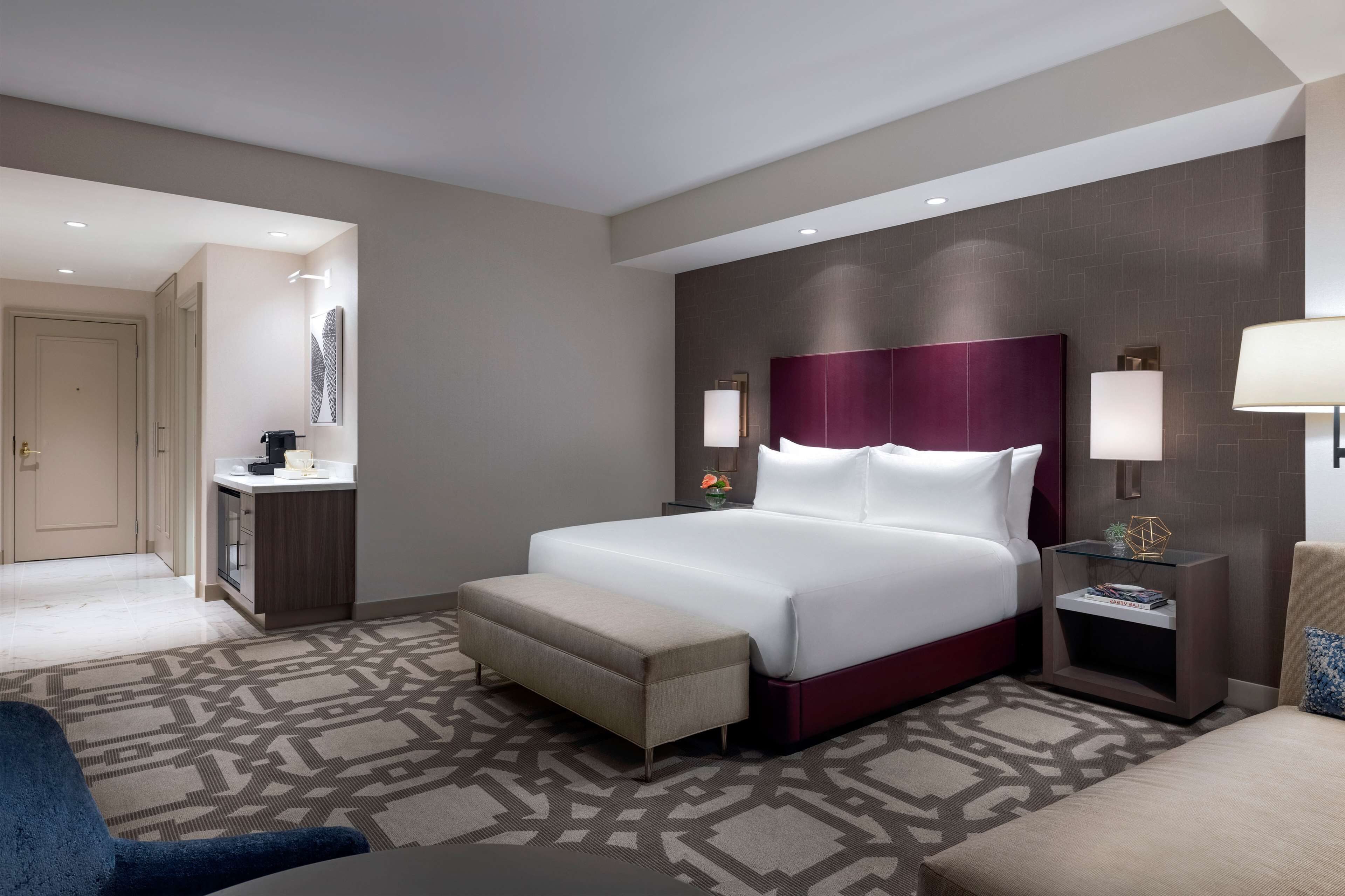 Una habitación de hotel con una cama grande, una mesita de noche con lámpara, una pequeña mesa con lámpara, un banco y un sofá.