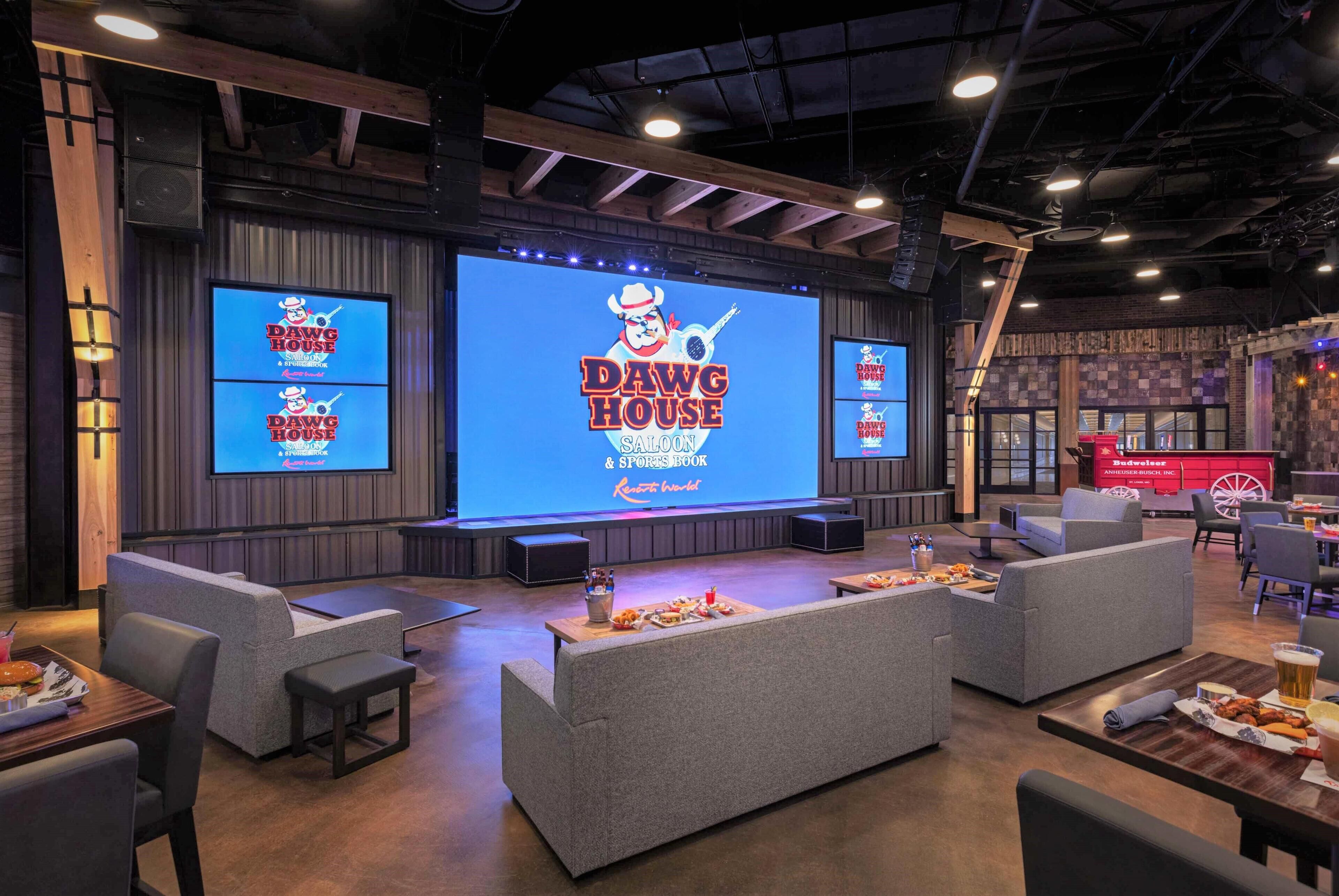 A modern lounge area with comfortable seating, a large screen displaying 'DAWG HOUSE' branding, and a bar area with food and drinks.