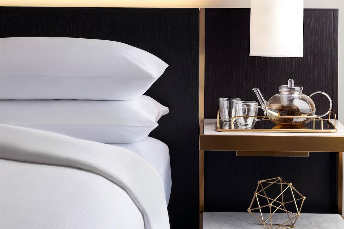 Una cama bien tendida con sábanas blancas, una mesita de noche con un juego de té y una escultura geométrica.
