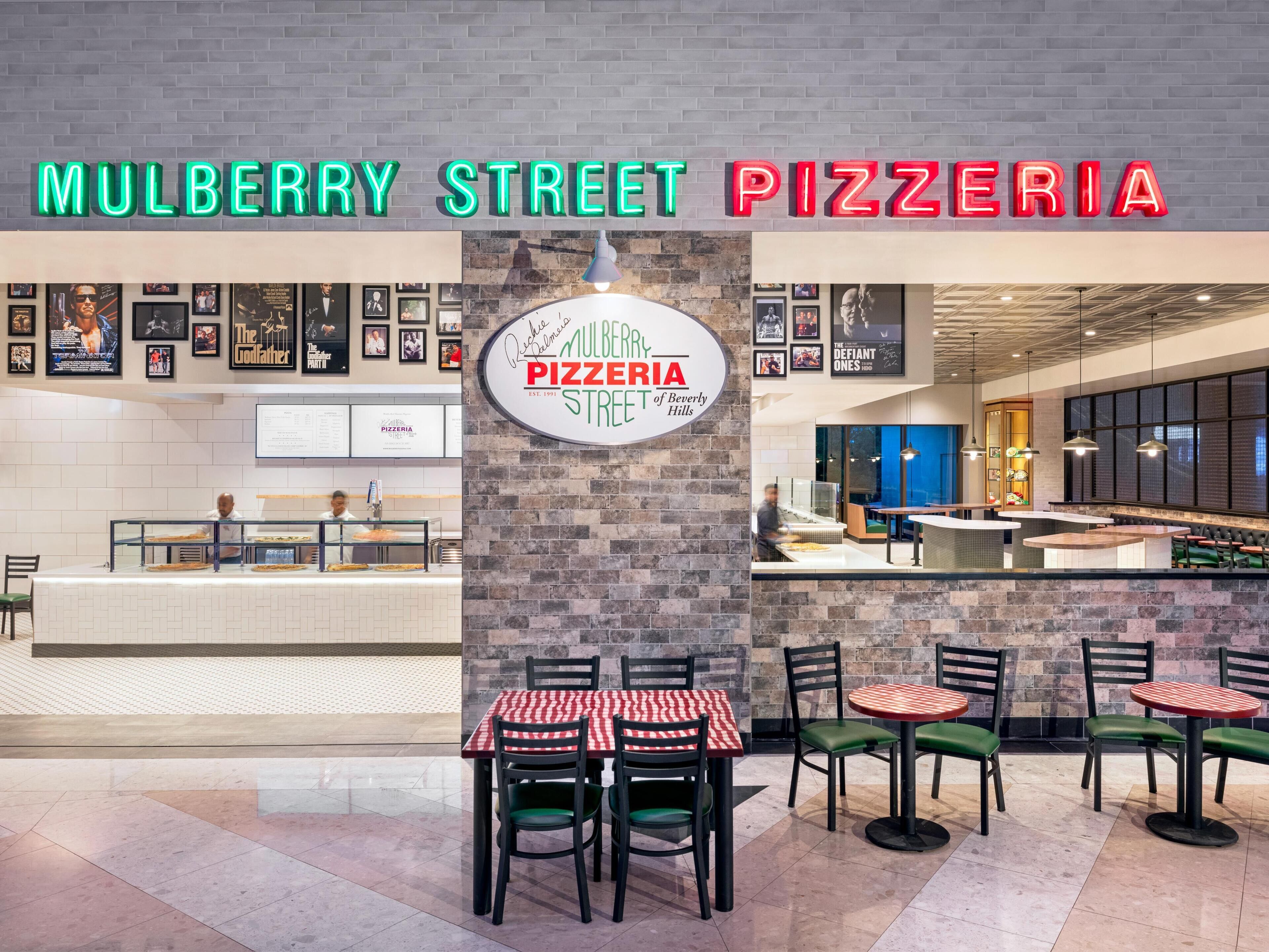 A pizzeria with a brick wall and a neon sign. There are tables and chairs for dining.