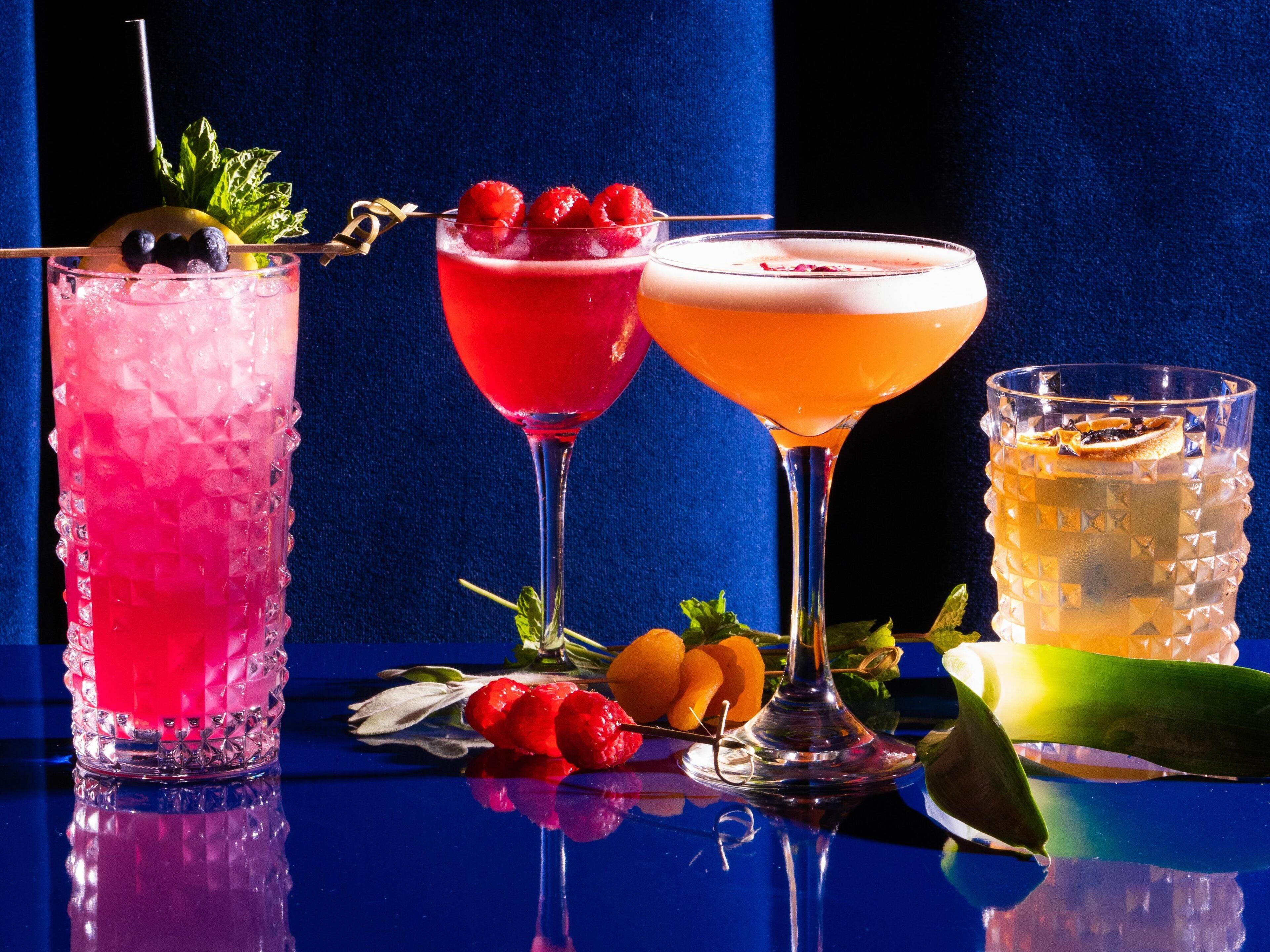 Three cocktails with garnishes and fruits on a reflective surface against a dark background.