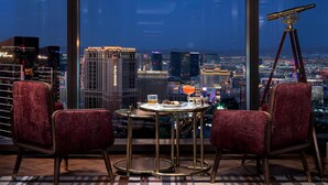 Une chambre située dans un immeuble de grande hauteur, offrant une vue panoramique sur la ville, avec deux fauteuils rouges confortables, une petite table ronde sur laquelle se trouve une boisson, et un télescope.