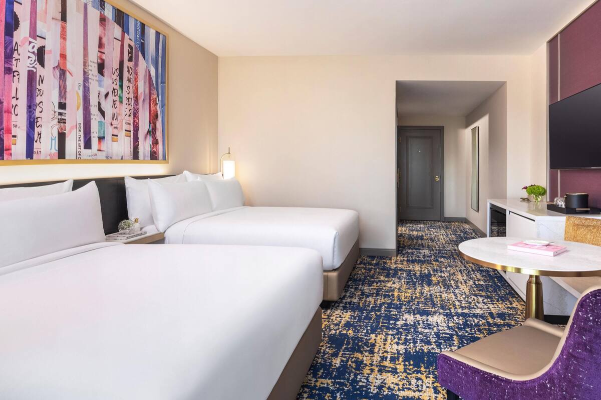 Habitación de hotel con dos camas, una mesa redonda, una silla morada, un televisor y un cuadro abstracto grande en la pared.