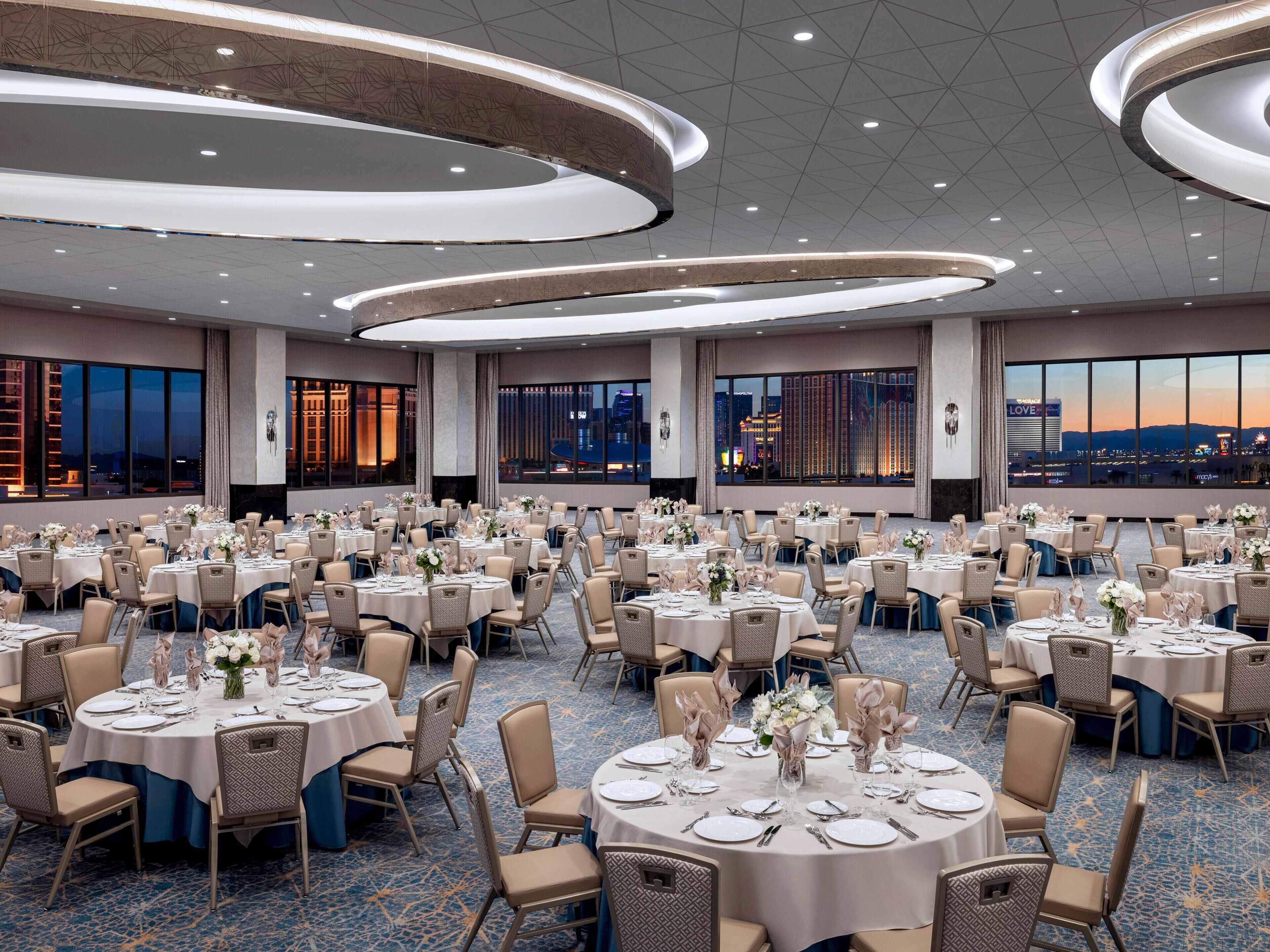 A large banquet hall with round tables set for dining, featuring a cityscape view through large windows.